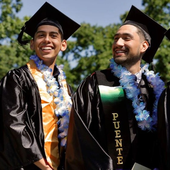 Two male Puente students dressed in graduation attire smiling at each other.